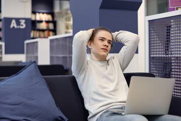 Sad unhappy college student in casual sweater holding hands behind head, feeling frustrated as he has to work on his diploma project, sitting on comfortable couch with open laptop on his lap