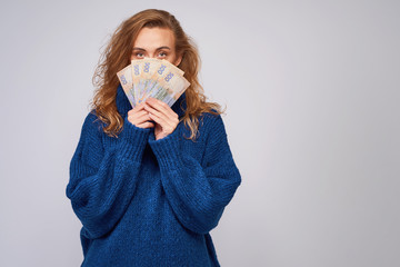 A cheerful woman in a sweater, covers part of the face with money hryvnia holding them in her hands on a gray background