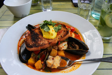 Bouillabaisse with toast served in a white bowl on a rustic table surrounded by glasses and a mojito with a cell phone laying nearby