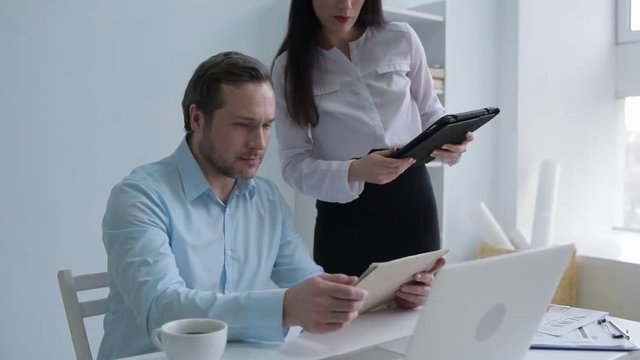 Office solicitation. Young lady with a tablet computer undoing buttons on her blouse and sitting on a table of her boss while flirting with him at work.