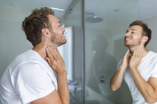 Man putting after shave perfume lotion or skin care cream for sensitive skin after shaving beard looking in bathroom mirror on neck. Male beauty, beard care skincare concept.