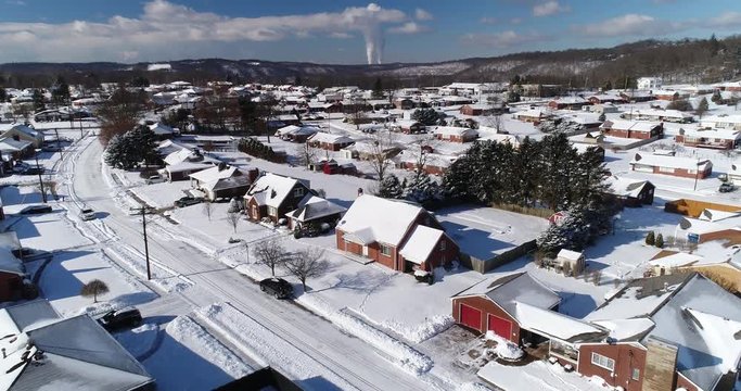A slow rising forward winter aerial establishing shot of snow covered roads and homes in a rust belt residential neighborhood. Factory smokestacks in the distance. Pittsburgh suburbs.  	