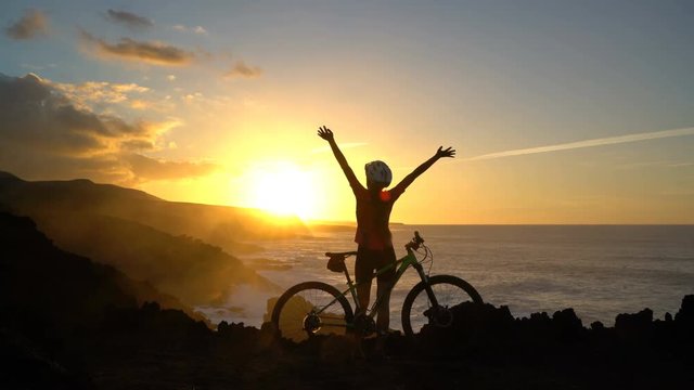 Carefree female MTB cyclist mountain biking raising hands by sea during sunset. Fit woman in sportswear is standing with bicycle on rock. Happy lady is enjoying nature on her bike.