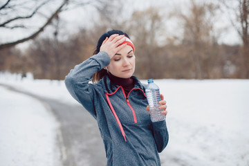 Portrait of Caucasian young woman tired after jogging outside in winter park. Girl drinking water when running outdoors. Exercising person female in street.