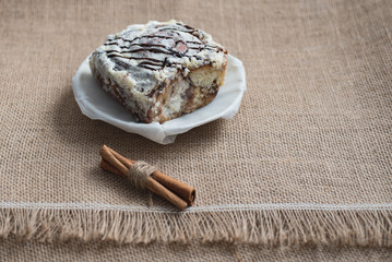 Still life of culinary spices and sweets. A sweet cake on a white porcelain plate with cinnamon sticks on a brown sackcloth surface in a cafe