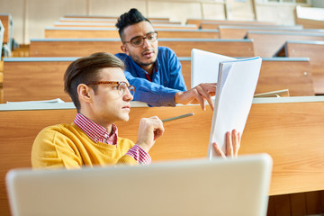 Low angle view of two young men, one of them Middle-eastern sitting at desks in modern lecture hall...