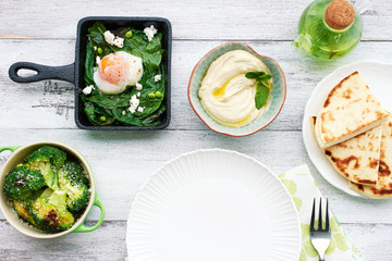 Top view of green shakshuka with spinach, basil and feta in a pan, baked broccoli, hummus and pita bread, green mint tea on white table with empty plate. Oriental style breakfast or lunch.