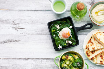 Top view of poached egg with spinach, basil and feta in a pan, baked broccoli with parmesan, flatbread and hummus, green tea on white table with copy space. Oriental style healthy breakfast or lunch.