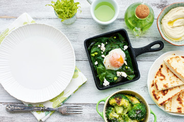 Top view of green shakshuka with spinach, basil and feta in a pan, baked broccoli, hummus and pita bread, green mint tea on white table with empty plate. Oriental style breakfast or lunch.