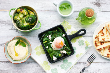 Poached egg with spinach, basil and feta in a pan, baked broccoli, hummus, flatbread and green tea on white table. Healthy oriental style breakfast or dinner. 