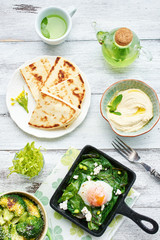 Poached egg with spinach, basil and feta in a pan, baked broccoli, hummus, flatbread and green tea on white table. Healthy oriental style breakfast or dinner. 