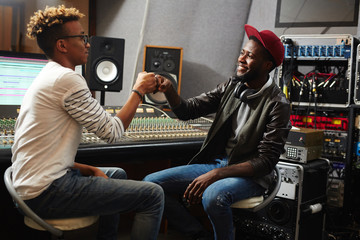 Two happy young musicians making punch bump while working in studio of sound recording