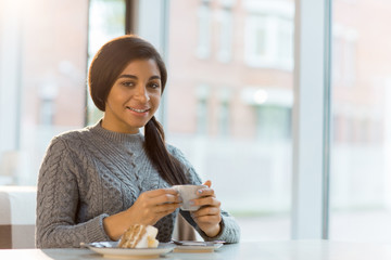 Obraz premium Young woman with cup of coffee and cake looking at camera while sitting in cafe