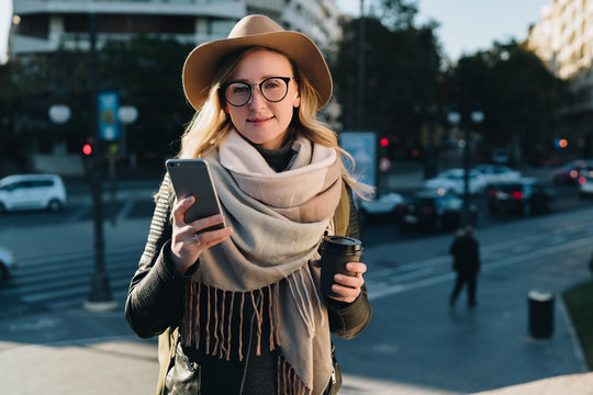 Sunny autumn day. Young attractive woman travels in hat and eyeglasses stands on city street, uses smartphone and drinks coffee. Hipster girl is looking for road. Vacation, adventure, trip.