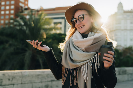 Sunny autumn day, backlight. Young attractive woman tourist in hat, eyeglasses and with backpack stands on city street,drinks coffee. Hipster girl walks, looks at sights. Vacation, adventure, trip.
