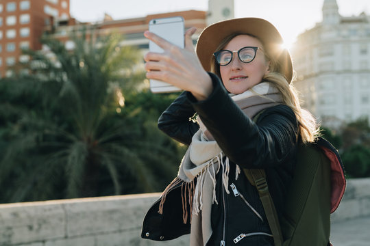 Sunny autumn day, backlight.Young attractive woman travels in hat, eyeglasses, with backpack stands on city street, uses smartphone, makes selfi.Hipster girl tourist walks. Vacation, adventure, trip.