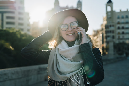 Sunny autumn day, backlight. Young attractive woman tourist in hat and eyeglasses stands on city street, talking on cell phone, smiling, laughing. Hipster girl walks. Vacation, adventure, trip.