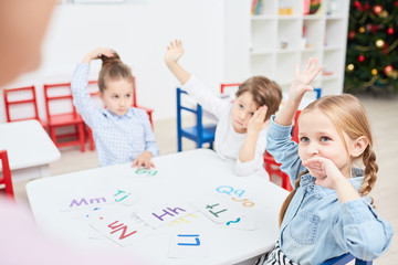 Cute kids by table raising their hands while learning alphabet with teacher