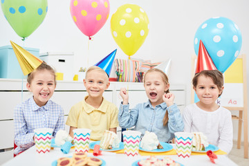 Excited kids in birthday caps sitting by festive table and having fun in kindergarten