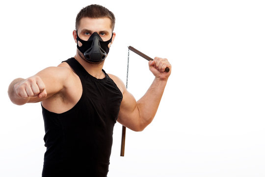 A young sporty blue-eyed man in a sporty black T-shirt, wearing boxing gloves and a black training mask is holding a metal nunchak in his hand on a white isolated background