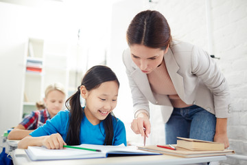 Teacher checking home assignment of cute schoolgirl and reading her notes in copybook