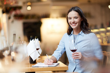 Happy young sommelier with glass of red wine tasting it and making notes - obrazy, fototapety, plakaty