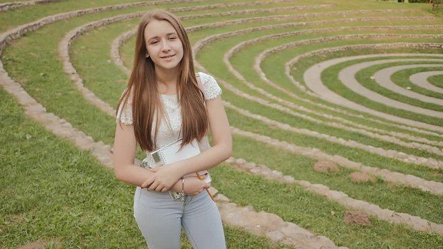 Beautiful young girl student with textbook and notes, smiling, standing in big green stadium. Rest during study.