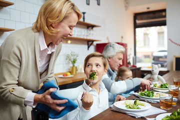 Little boy eating steamed broccoli and looking at his grandmother during dinner in the kitchen