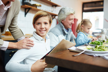 Cute boy with open book sitting by table during dinner and looking at camera