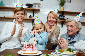 Happy family members clapping their hands while little girl going to blow candles on birthday cake