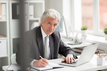 Experienced director in formalwear sitting by workplace, networking and making notes