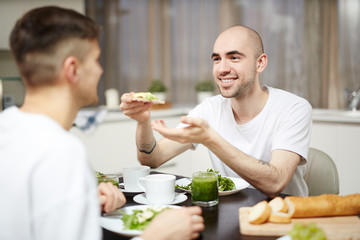 Fototapeta na wymiar Happy young man giving healthy sandwich to his partner during breakfast