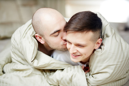 Happy young gay men wrapped in blanket lying in bed while spending time together