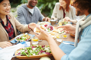 Young man passing plate with fresh salad and baked potatoes over served table to one of girls