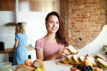 Happy woman with sandwich looking at camera while standing by served table with her friend on background