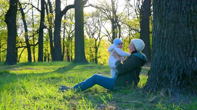 Mother plays with her baby in a spring park under a big oak tree.