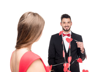 girlfriend standing near happy boyfriend bound with garland of hearts isolated on white, valentines day concept