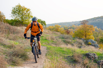 Naklejka na ściany i meble Cyclist in Orange Riding the Mountain Bike on the Autumn Rocky Trail. Extreme Sport and Enduro Biking Concept.