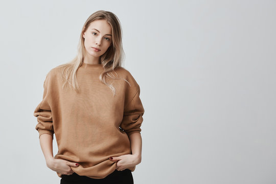 Cute pretty beautiful woman wears loose sweater and black trousers, holds her hands in pocket, looks with appeal at camera. Pleasant-looking girl posing against gray background
