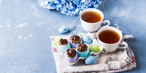 Easter sweet cup cakes treets in colorful pastel egg shells with chocolate and hydrangea flowers on blue background. Festive holiday home made food with cups of tea