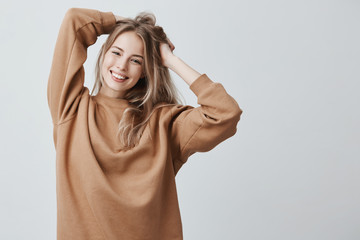 Beautiful pretty charming young blonde woman in loose sweater smiling happily, having fun indoors,...