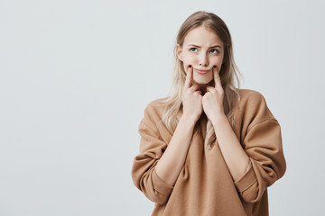 Indoor shot of pretty blonde woman making grimace, touching her cheeks with fingers, looking upwards, having displeased expression. Face expression and negative emotions - obrazy, fototapety, plakaty