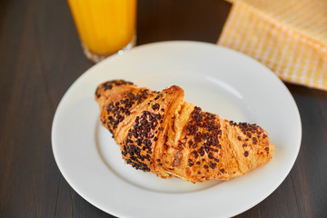 Fresh croissant on a ceramic plate and kitchen towel with a cup of orange juice. Freshly baked croissant on a dark wooden table.
