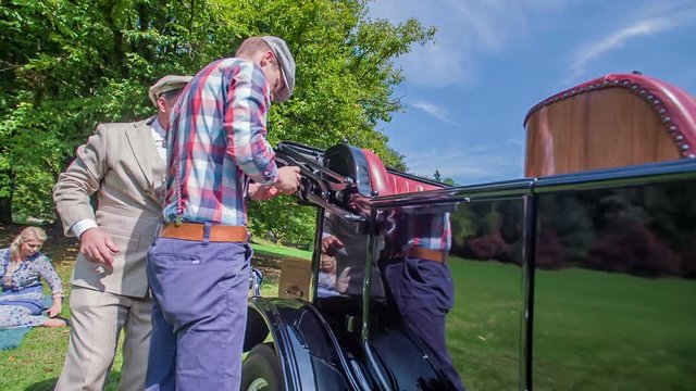 A young man is trying to tie the roof to a vintage car. The driver comes and wants to help him out. A young woman is sitting on a blanket nearby.
