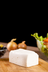 Close up on feta cheese lying on wooden board next to onions, fresh salad and glass bowl of olives on black background