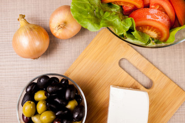 Close up top view of table with feta cheese on wooden board, onions and fresh salad with tomatoes next to a glass bowl with olives
