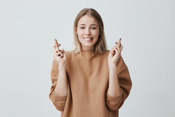 Portrait of wishful young woman in casual clothes with blonde dyed hair, crossing her fingers,...
