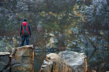 Traveler stands on granite stones amidst a calm lake. Back view