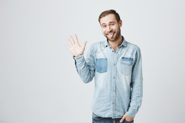 Handsome positive young bearded man in stylish clothes raising hand and gesturing as if saying hello, having good mood and lots of fun, sticking out his tongue. Attractive male waving to his friends