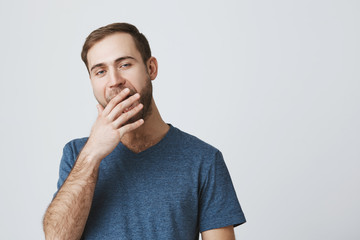 Sleepy muscular guy with dark hair and beard is tired work, yawning, opening widely his mouth, hiding his opened mouth behind his hand. Bored and annoyed european male model